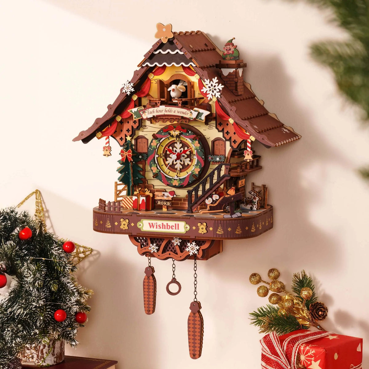 ROKR LCB01 Christmas cuckoo clock mounted on a wall beside a decorated Christmas tree and wrapped gift.