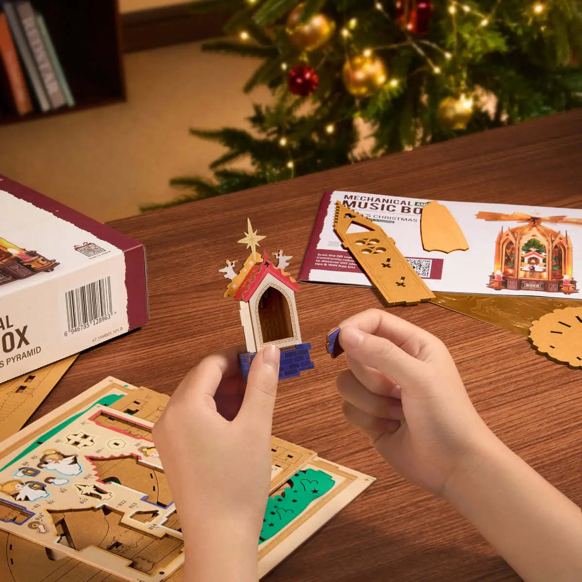 Hands assembling a ROKR AMB01 wooden component beside puzzle sheets, illustrated instructions and the kit box